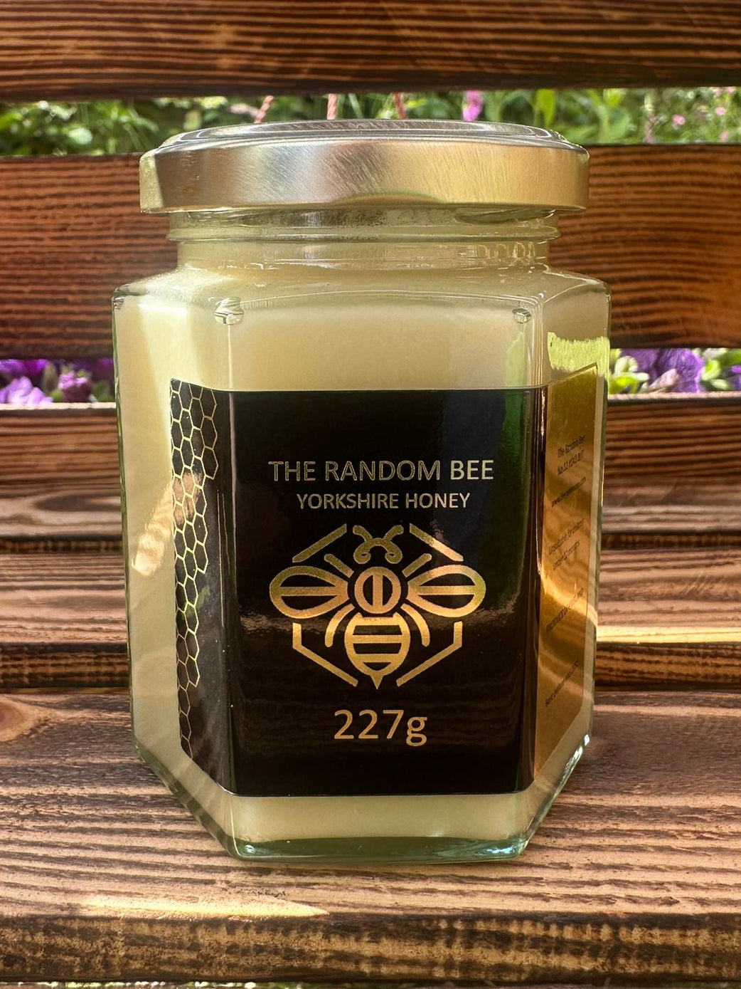 Jar of 227g creamed honey from 'The Random Bee' on a wooden surface with a blurred natural background