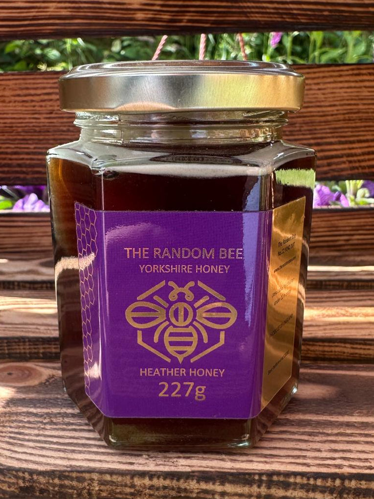 Jar of 'The Random Bee' 227g Yorkshire Heather Honey on a wooden surface with blurred greenery in the background.