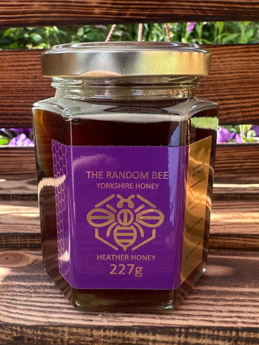 Jar of 'The Random Bee' 227g Yorkshire Heather Honey on a wooden surface with blurred greenery in the background.